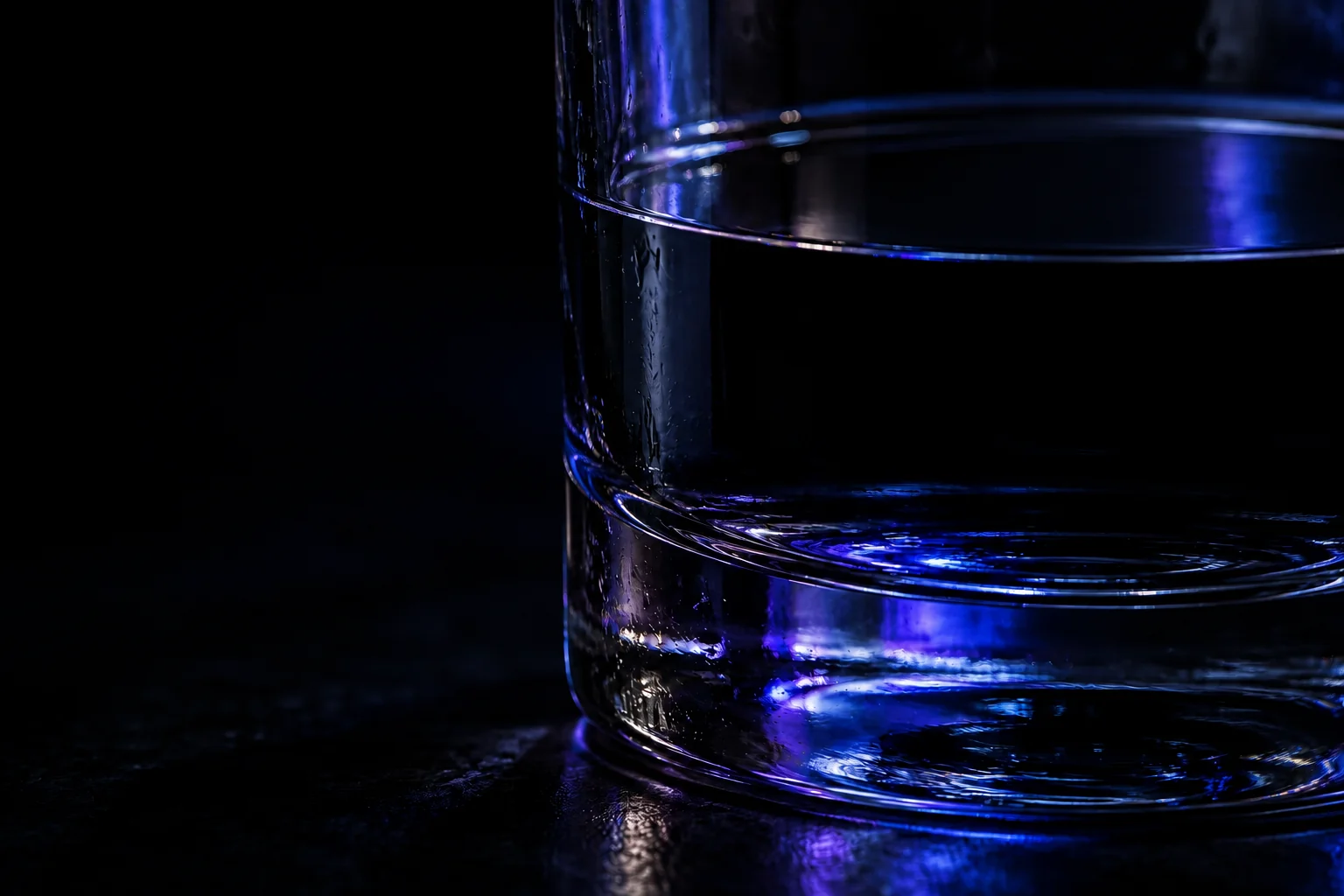 A glass of dark water, perfectly still until a falling drop opens rings of blue light across its surface.